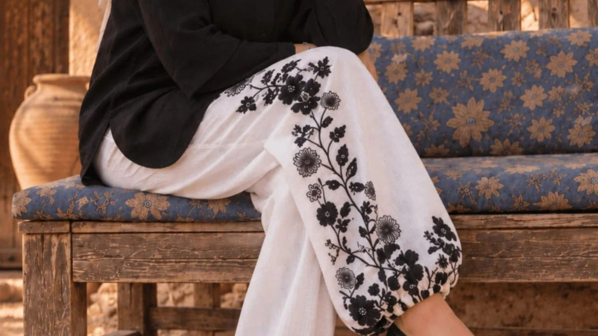 White wide-leg pants with black floral embroidery, worn by a model sitting on a rustic wooden bench with blue patterned cushions.